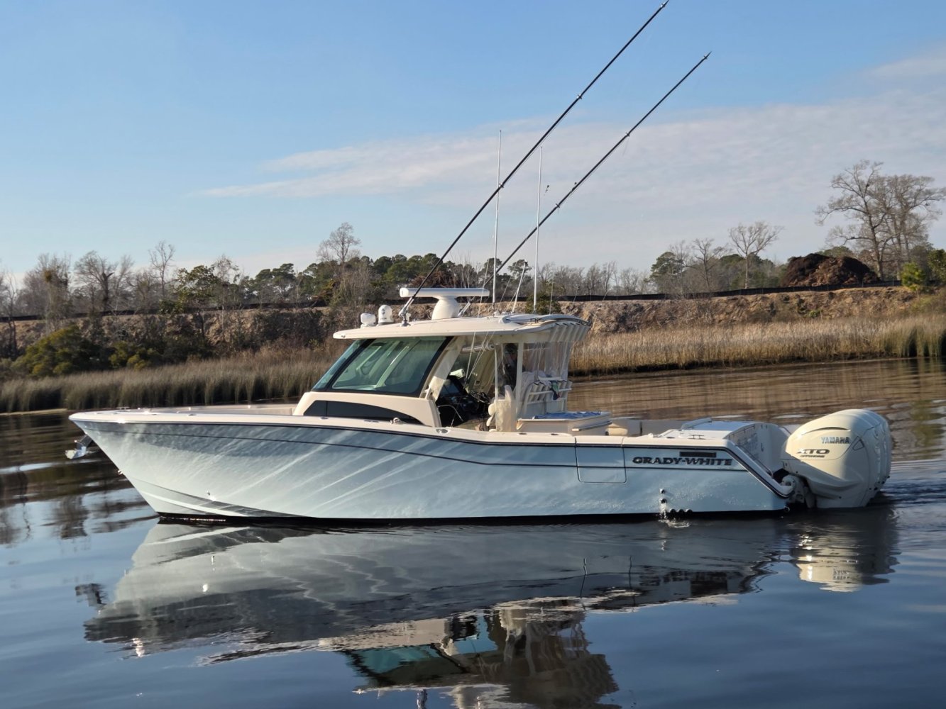 A fishing boat with rods and two engines sits on calm water near a grassy shoreline.
