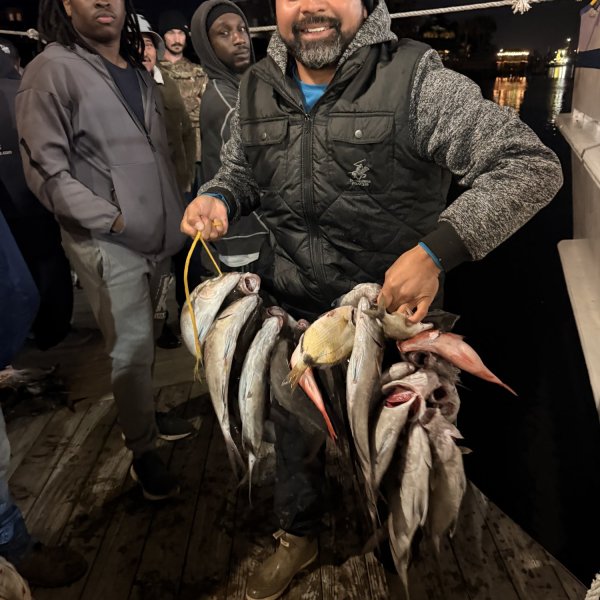 Smiling man on boat holding a string of fish at night, surrounded by others.