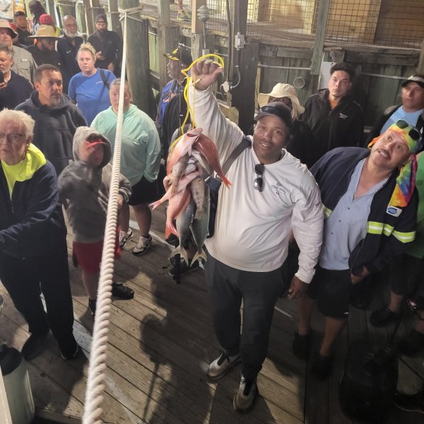 Man on dock holds up several fish, surrounded by onlookers.