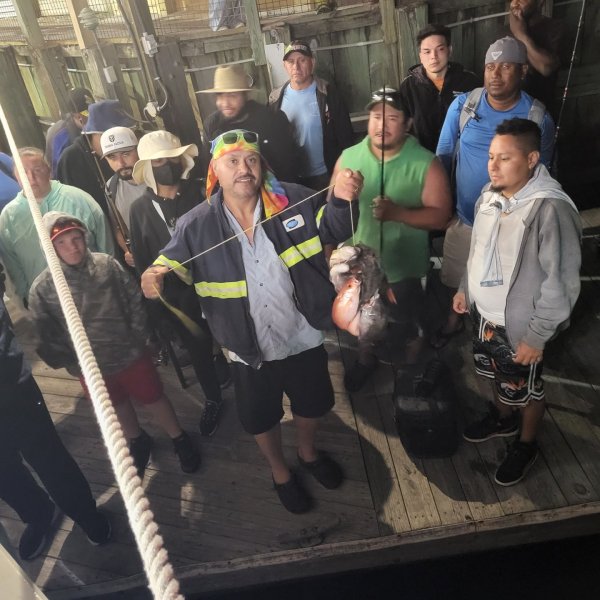 Man holding fish on dock with a group of people around him, some holding fishing rods.