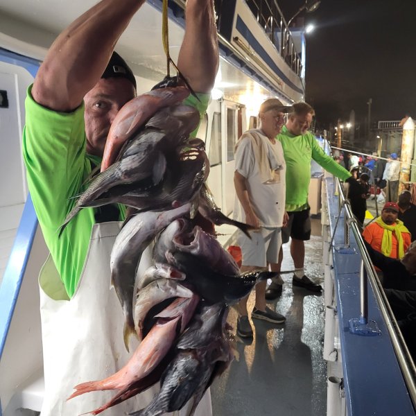 Person holding a bundle of fish on a dock, with others standing nearby, illuminated by boat lights at night.